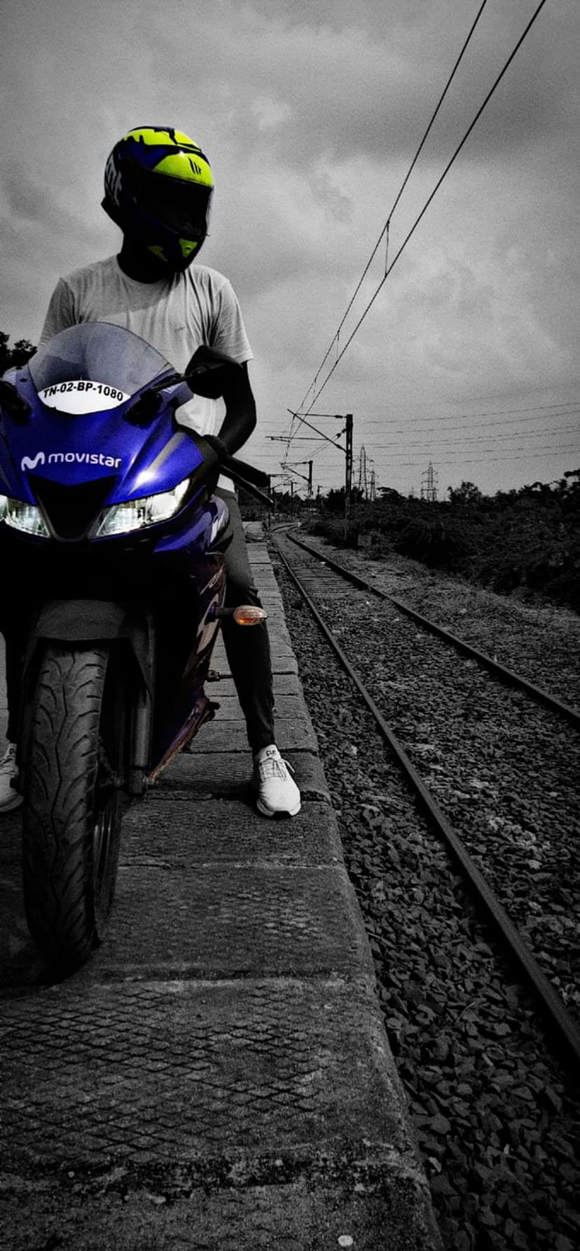 Motorcyclist in a yellow helmet sits on a blue motorcycle beside railway tracks under industrial power lines.