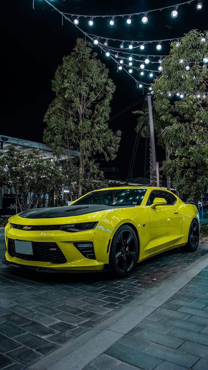 Yellow Chevrolet Camaro parked on a brick street at night under warm string lights and green leafy tree branches.