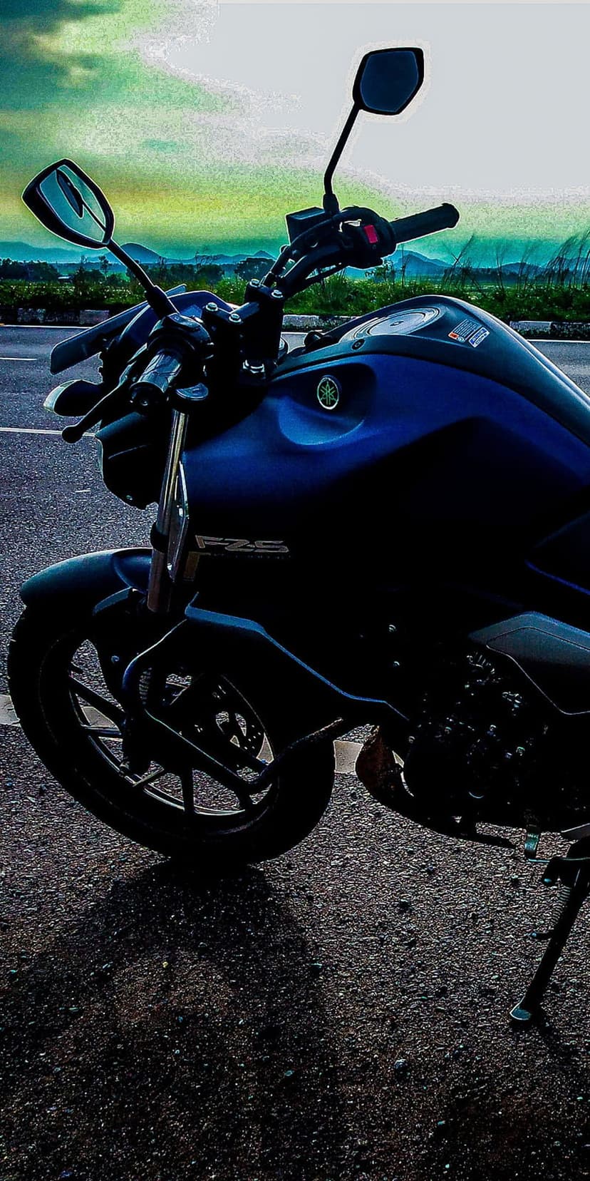 Blue Yamaha FZS motorcycle parked on a tarmac road with a blurred mountain range and dramatic sky in the background.