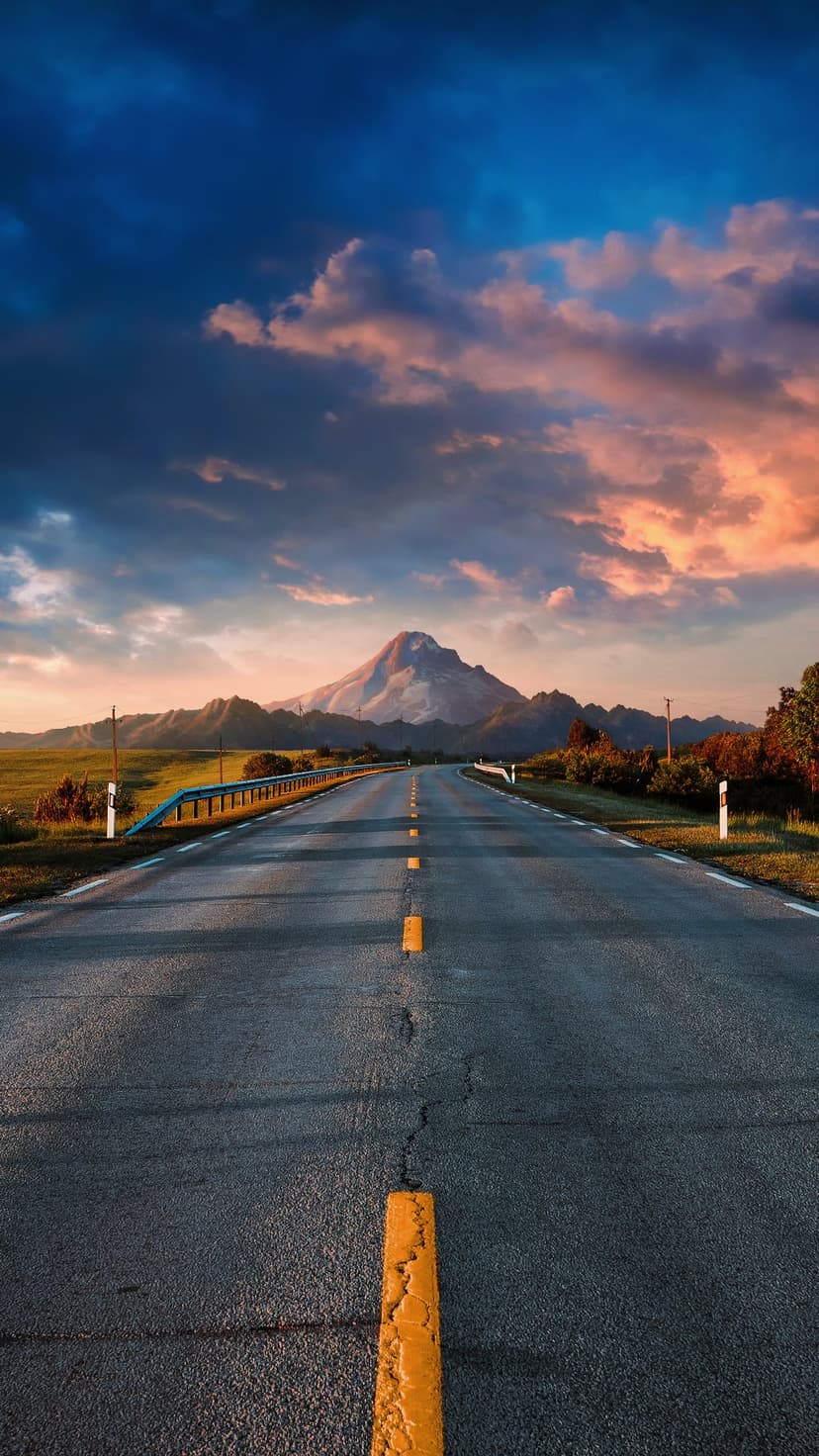 Asphalt road with yellow lines leading to a distant mountain peak under orange and blue sunset clouds.
