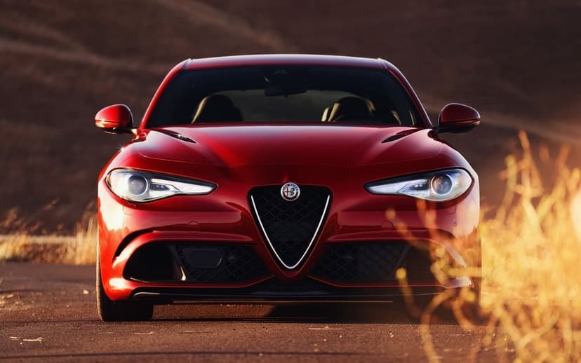 Red Alfa Romeo sports sedan centered on a road during golden hour with warm sunset lighting and blurred hills.