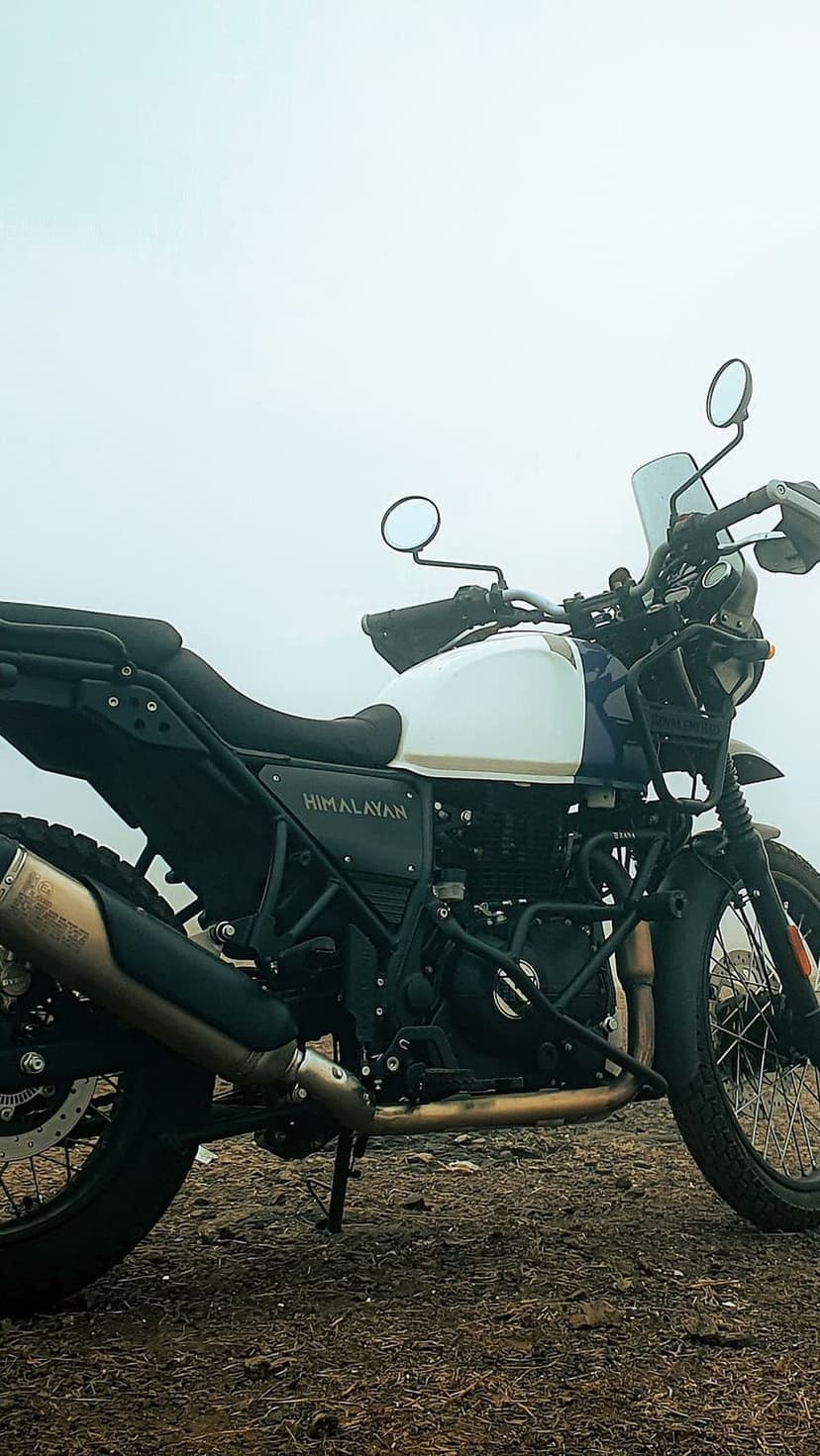 Royal Enfield Himalayan motorcycle with a white fuel tank parked on a dirt path under a foggy, overcast sky.