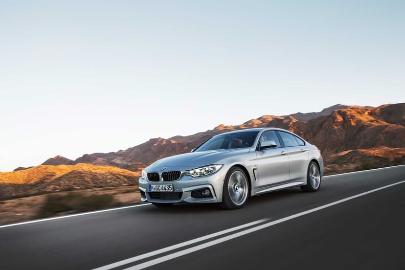 Silver BMW 4 Series Gran Coupe driving on a paved highway with rugged mountains under a clear sky at sunset.