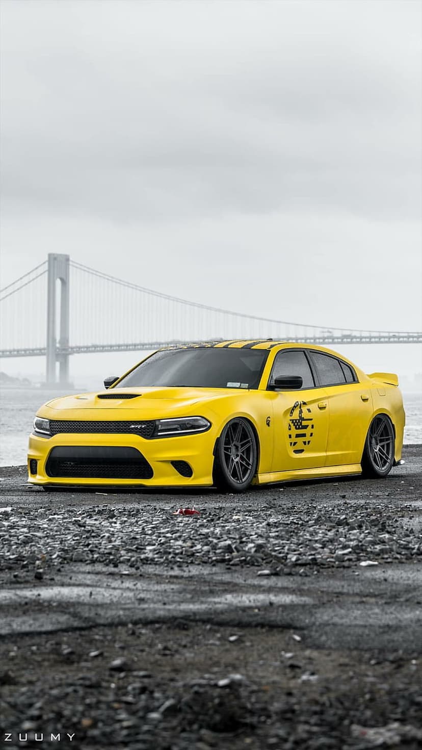 Yellow Dodge Charger Hellcat with custom wheels parked on rocks near a large bridge under cloudy skies.