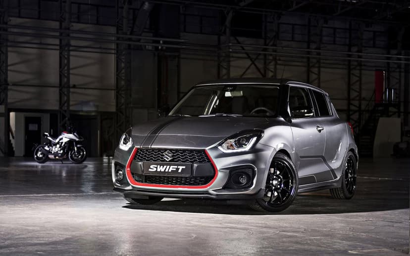 Silver Suzuki Swift Sport with red grille accents and black wheels in a dark industrial garage with a motorcycle.