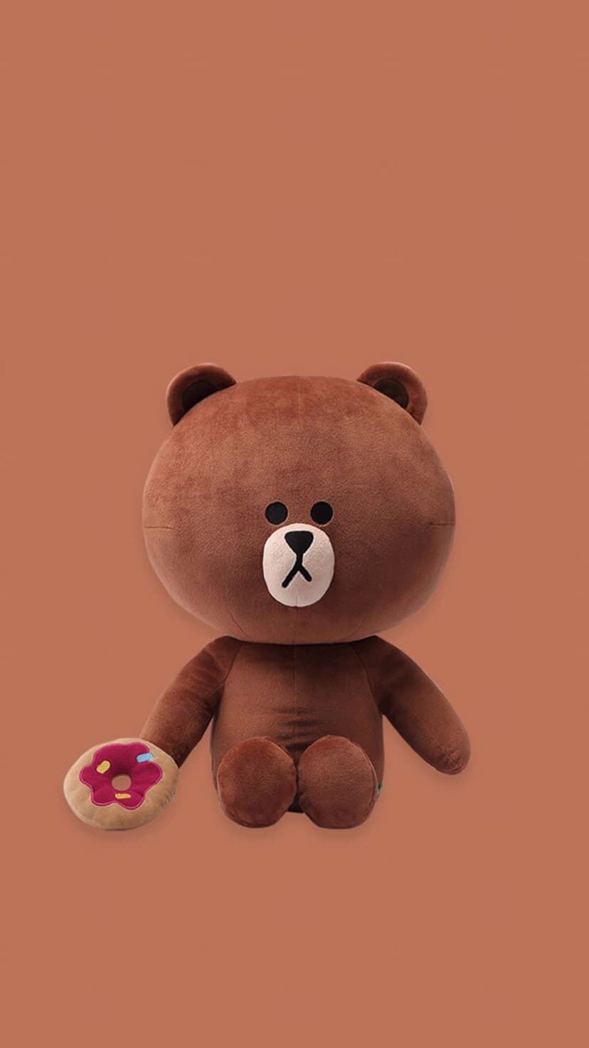 Brown bear plushie holds a pink frosted donut with sprinkles against a solid warm orange-brown background.