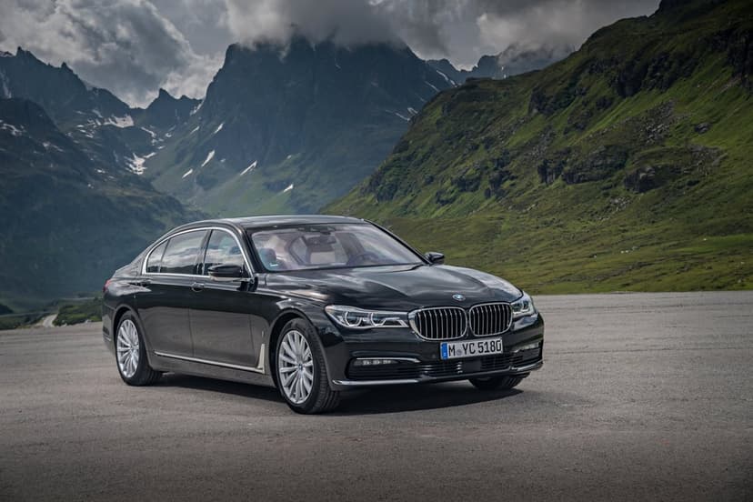 Black BMW 7 Series sedan parked on an asphalt lot against a backdrop of green slopes and jagged rocky mountains.