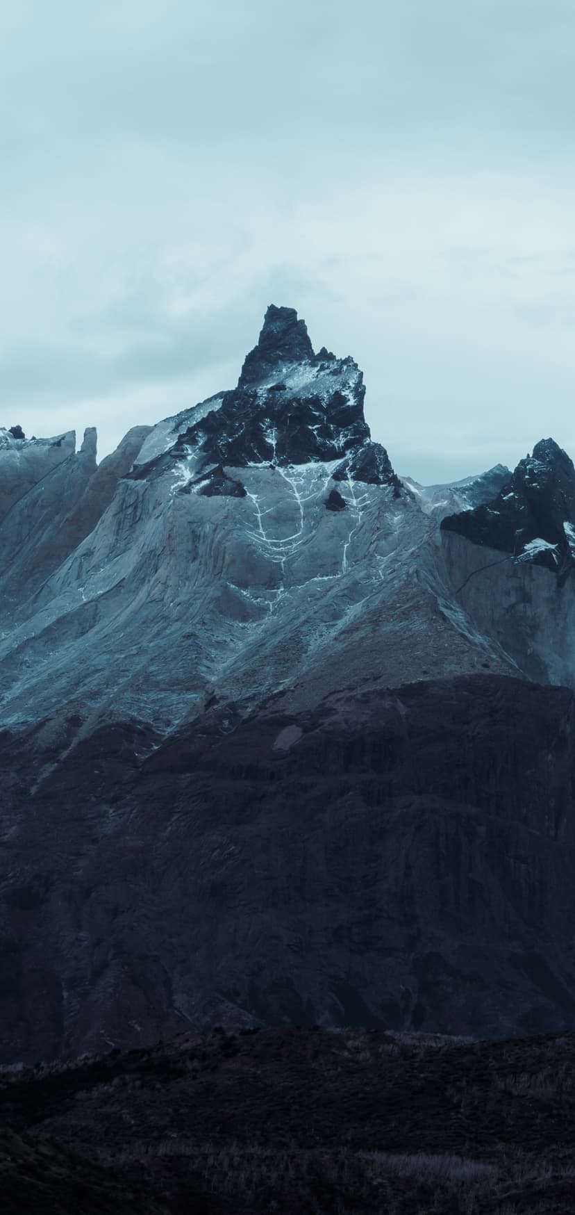 Sharp mountain peak with light snow dustings rises above dark forests under a heavy, overcast winter sky.