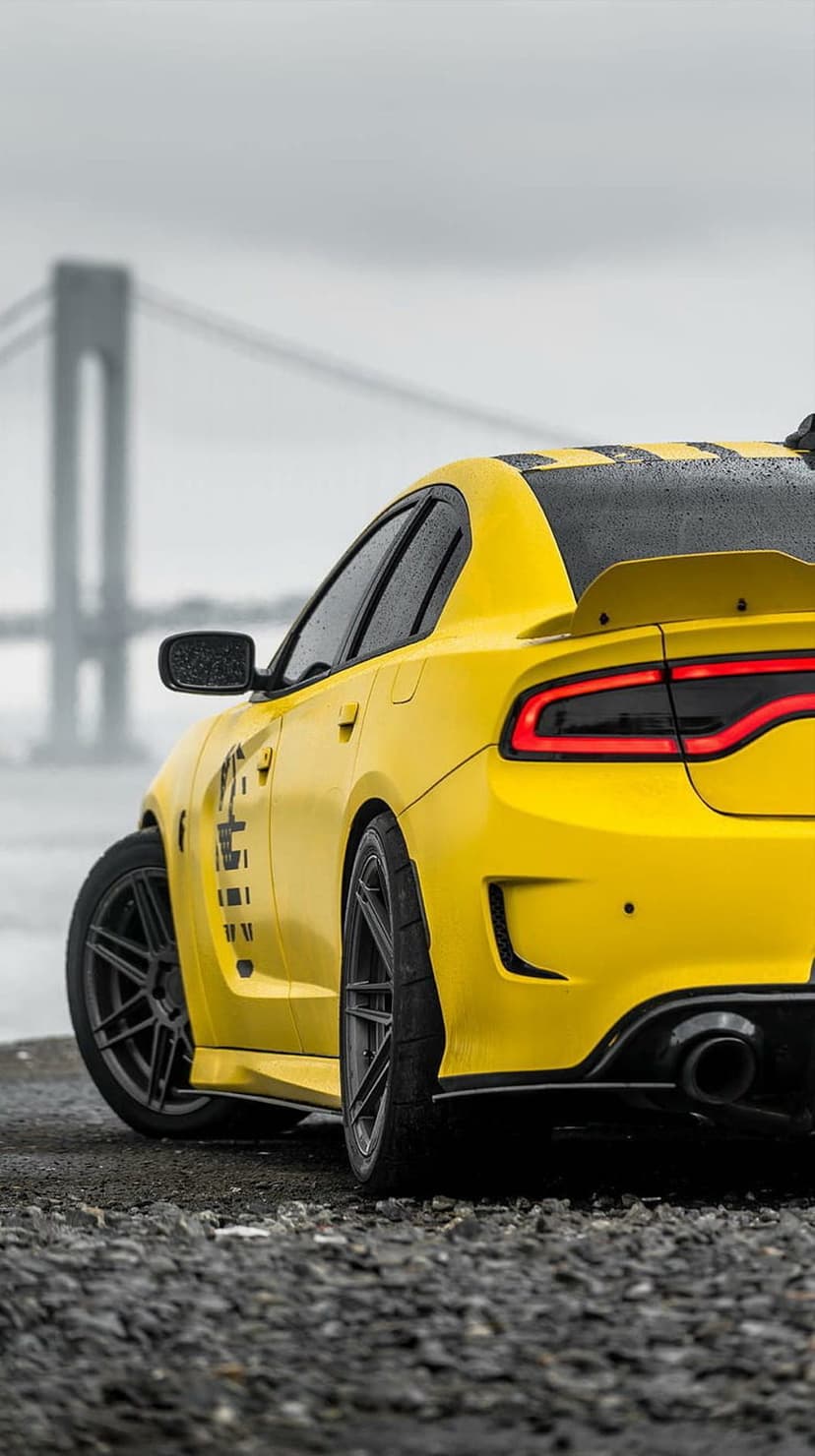 Yellow Dodge Charger with black stripes parked on wet gravel near a bridge during a rainy day with lit taillights.
