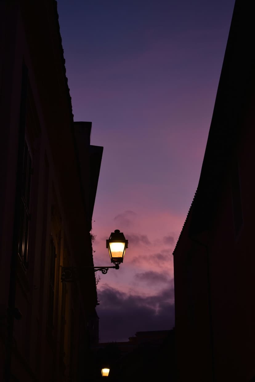Vintage street lamp glowing in a narrow alleyway under a vibrant purple and pink twilight sky with silhouettes.