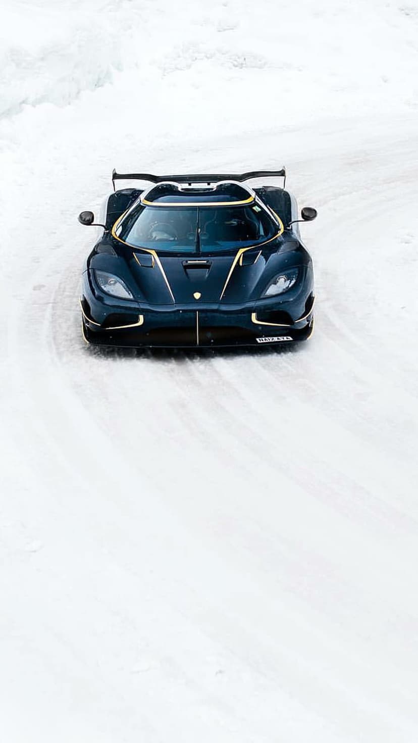 Dark blue Koenigsegg supercar with gold accents drifting on a snow-covered track seen from a high-angle view.