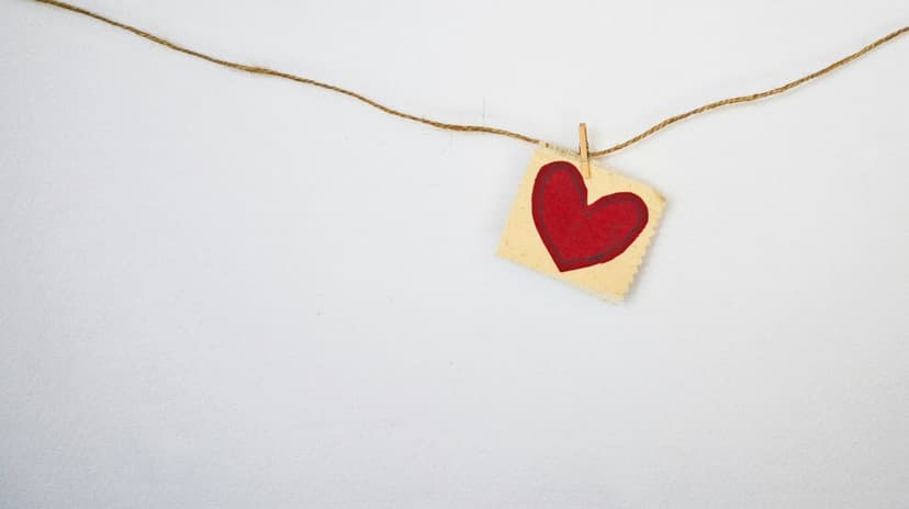 Red heart shape cut out of beige paper hanging from a wooden clothespin on a string against a white background.