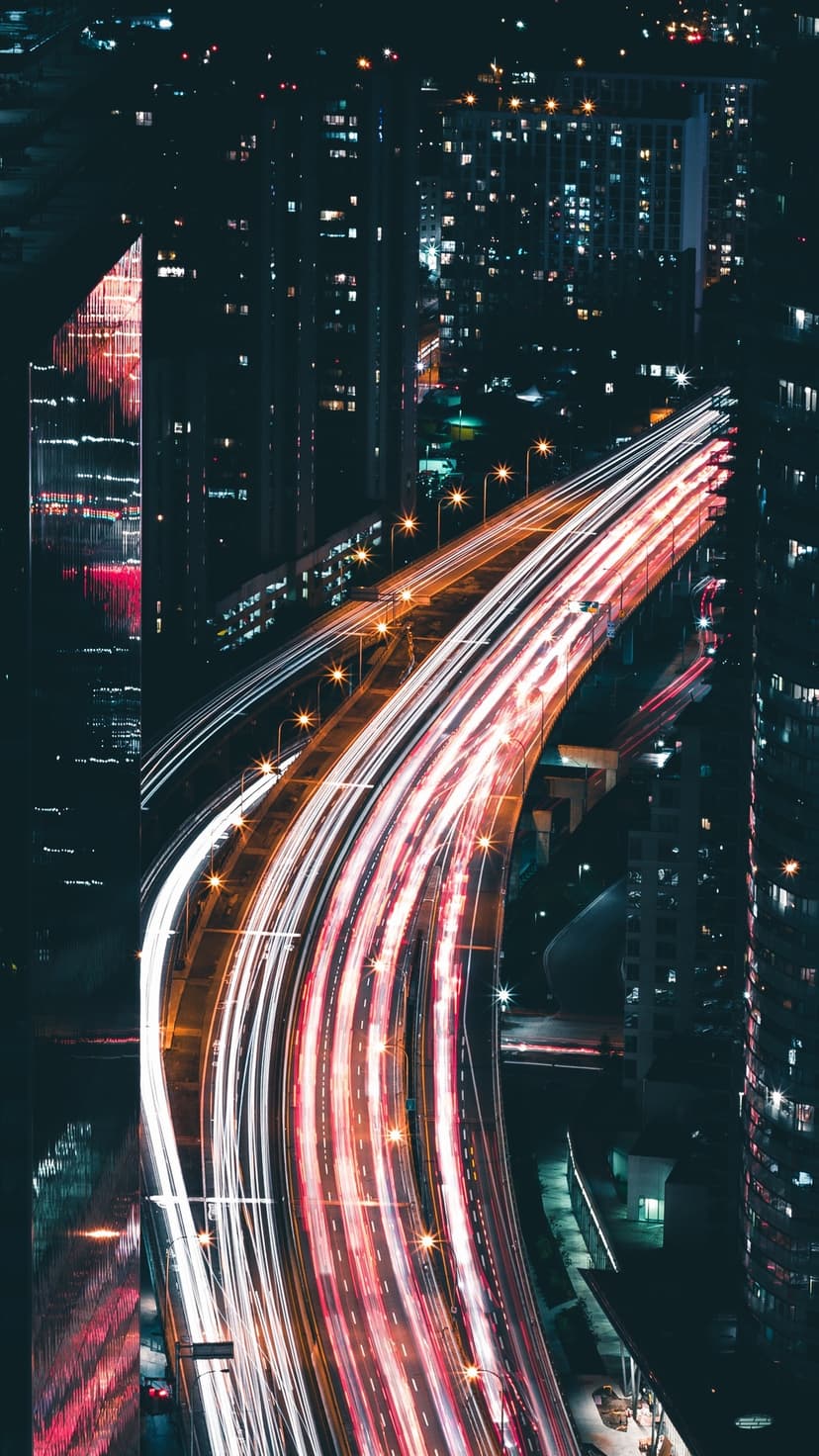 Glowing red and white car light trails wind between illuminated skyscrapers in a vertical city night scene.