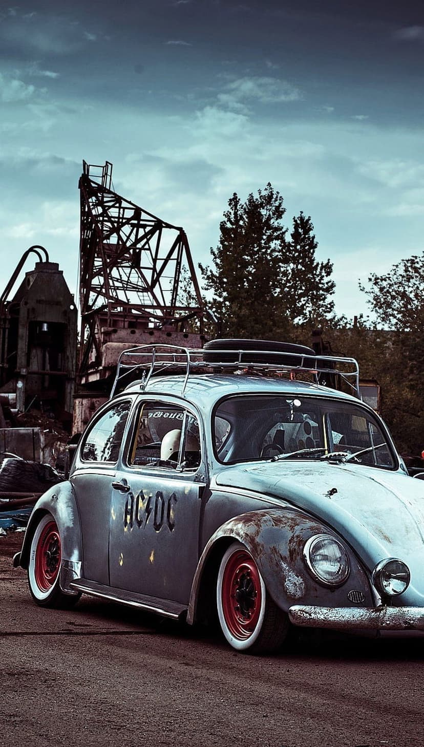 Rusty Volkswagen Beetle with AC/DC lettering parked near industrial gears under a moody, overcast sky.