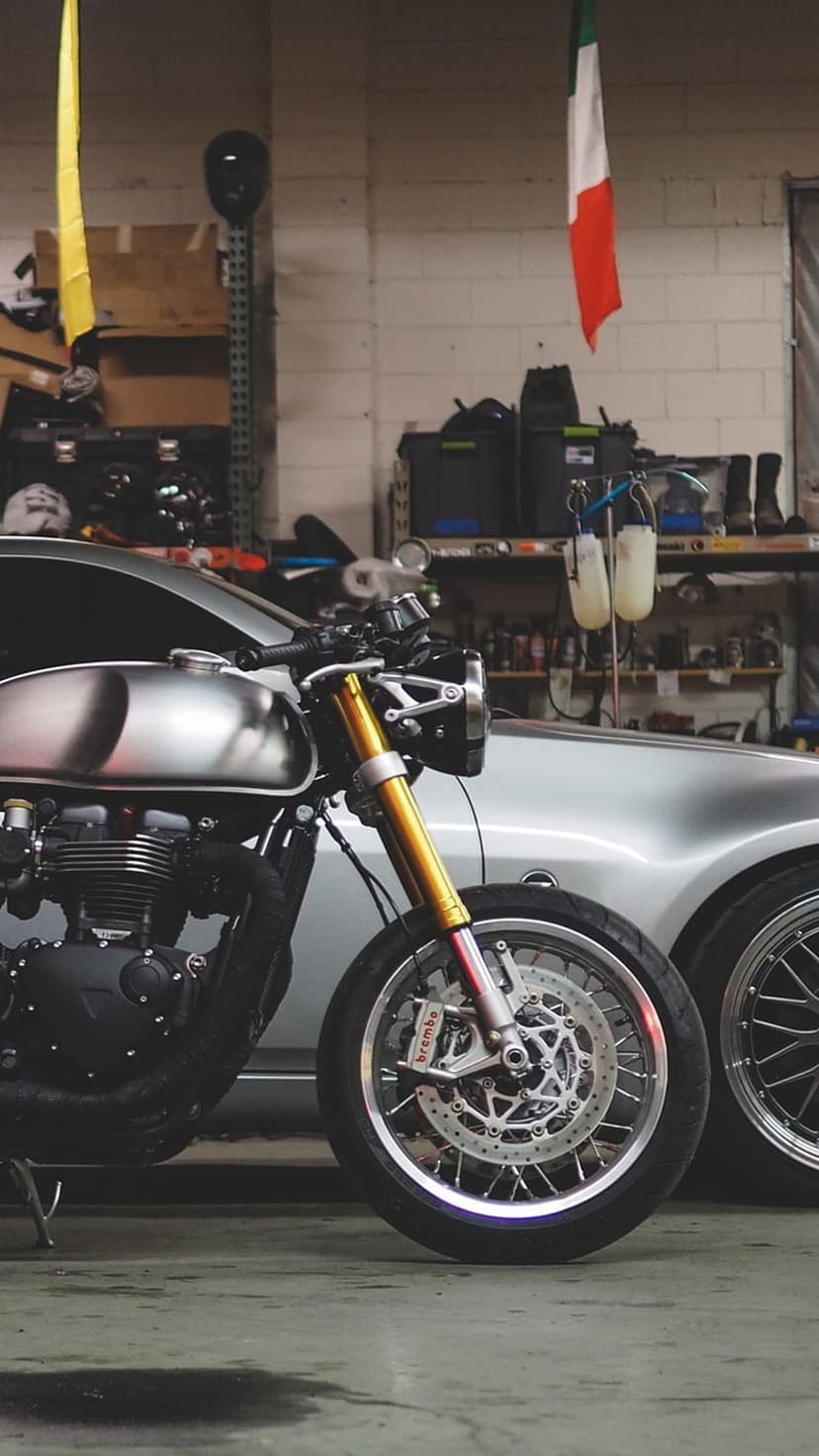 Silver cafe racer with gold front suspension forks parked next to a silver car in a professional garage.