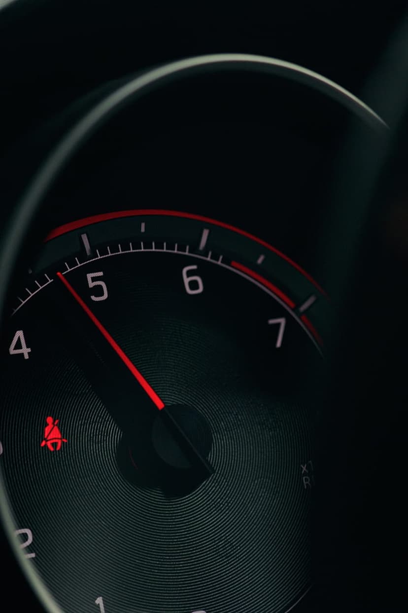 Red tachometer needle pointing to five thousand RPM beside a glowing seatbelt warning light on a dark dashboard.