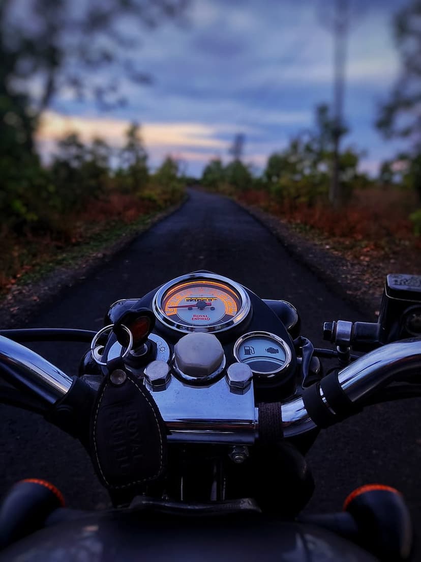 Motorcycle speedometer and fuel gauge glow on a chrome dashboard as a dark road winds through autumn trees at dusk.