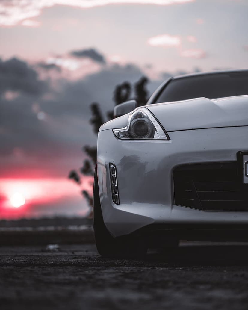 White sports car front grille and glowing headlights set against a dramatic pink and orange cloudy sunset sky.