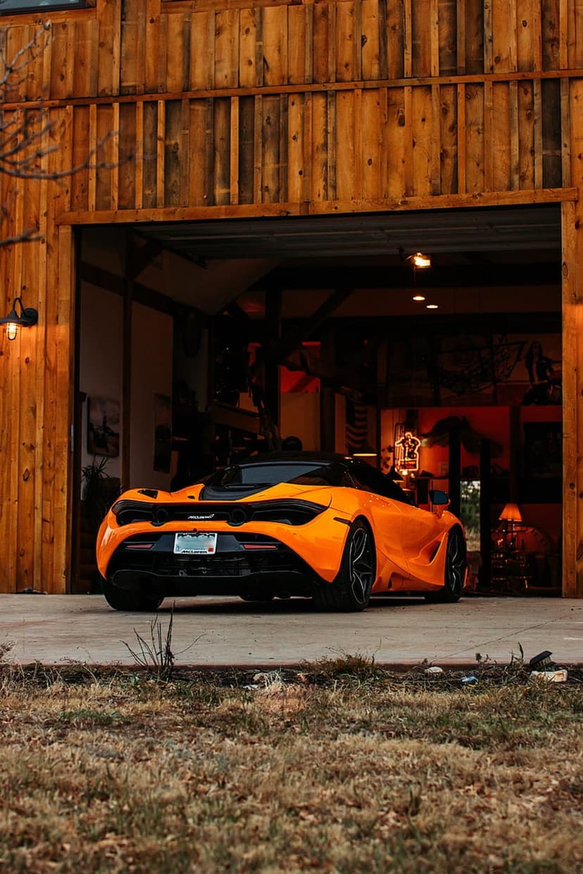 Orange McLaren sports car parked on a dark floor inside a rustic garage with vertical wooden wall planks.