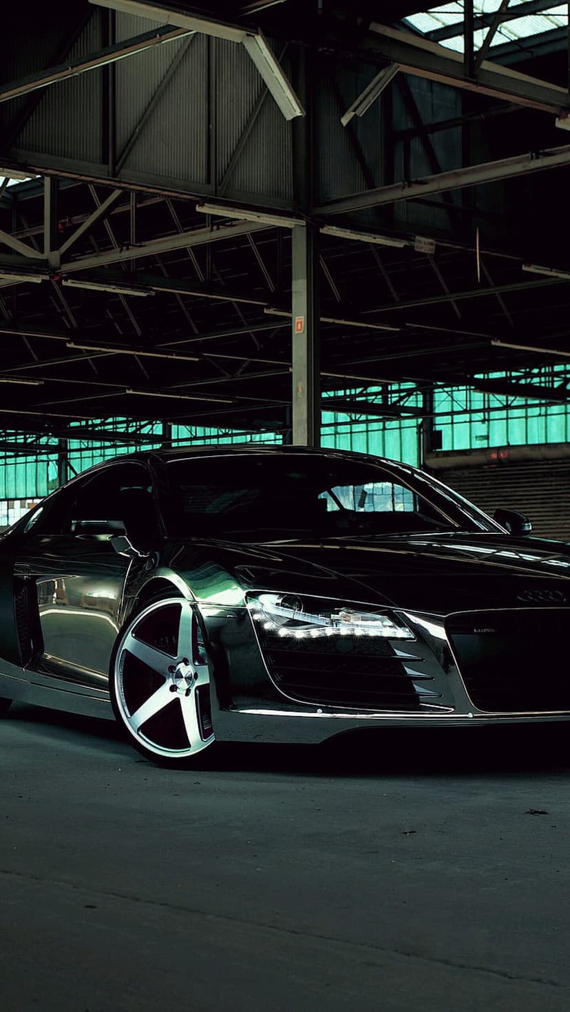 Black Audi R8 supercar with glowing LED headlights parked in a dark warehouse with green tinted windows.