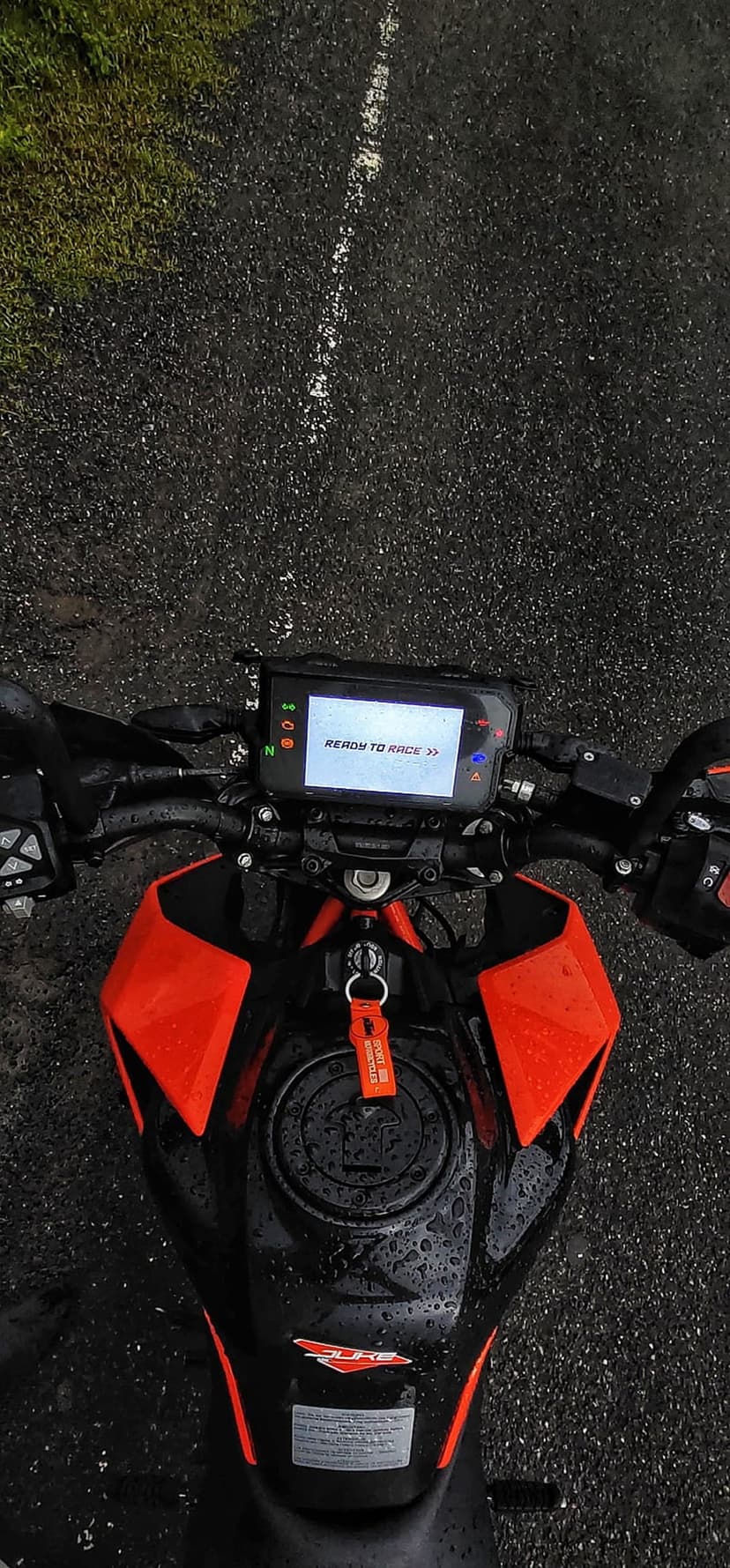 KTM motorcycle dashboard displays Ready to Race text on a wet asphalt road with raindrops on orange bodywork.