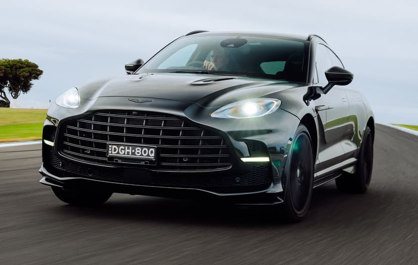 Deep green Aston Martin DBX707 luxury SUV with lit headlights speeding on a paved racetrack circuit.