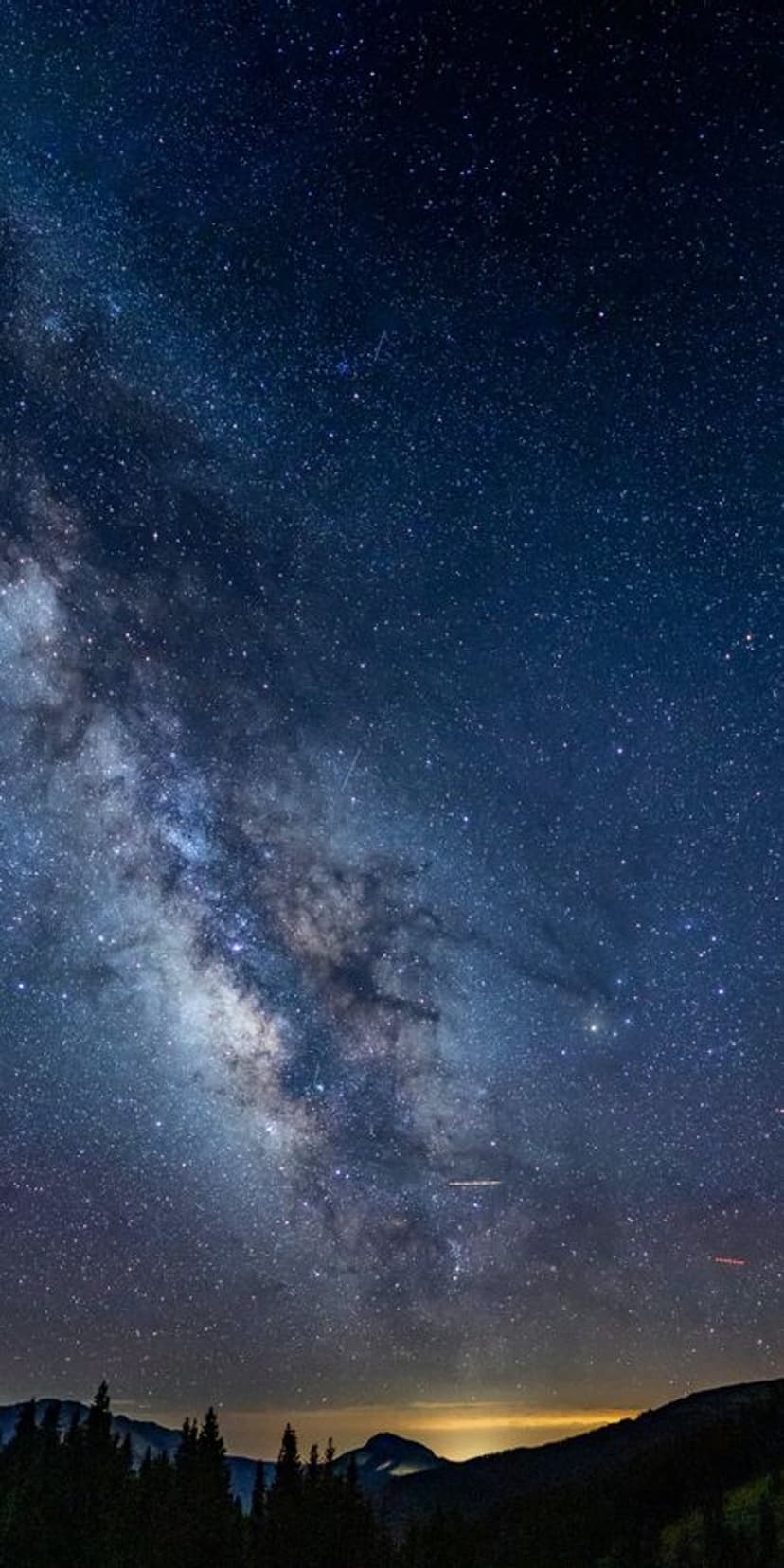 The Milky Way galaxy arches over dark mountain peaks and forest silhouettes against a starry night sky.
