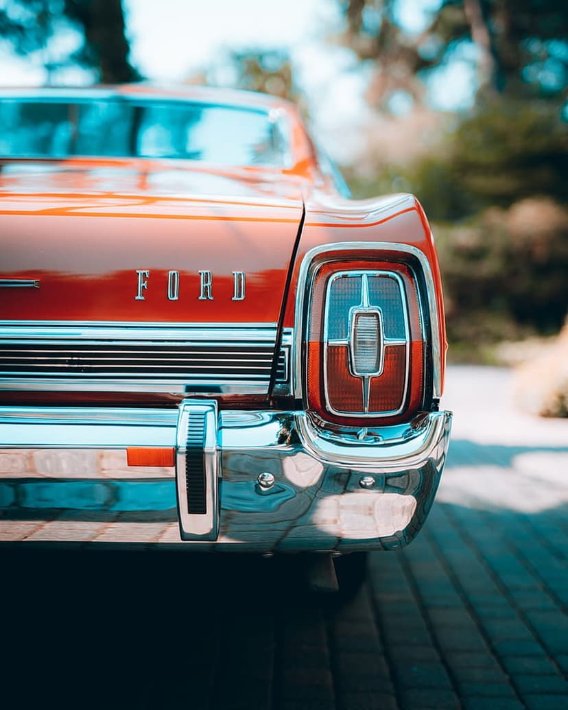 Red Ford vintage car rear with a chrome bumper, intricate taillight, and metallic lettering against green trees.