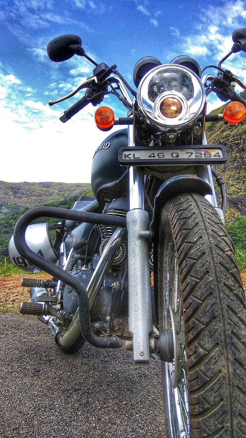 Royal Enfield motorcycle on a gravel mountain road under a clear sky with lush green hills in the background.
