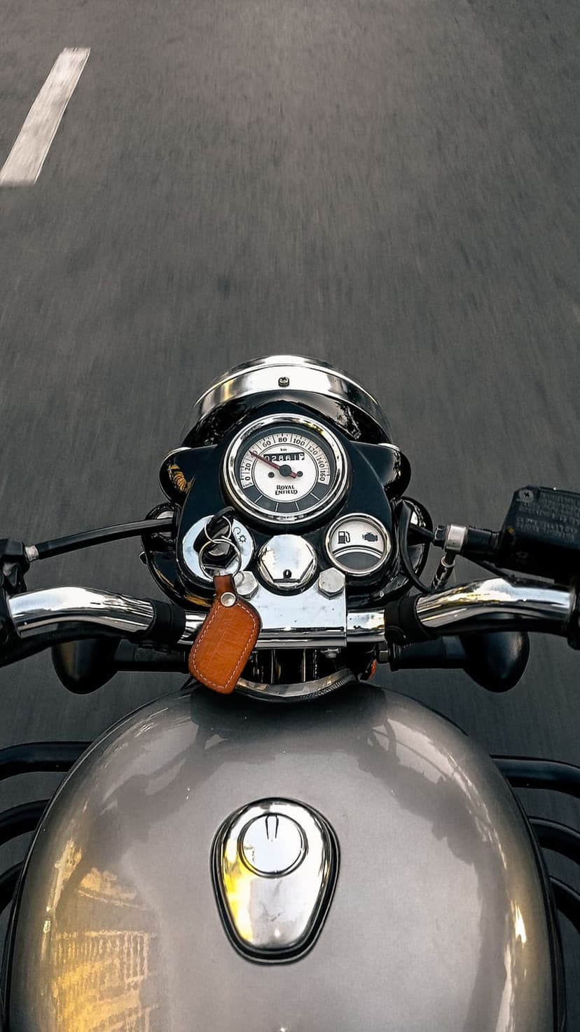 Royal Enfield handlebars and chrome speedometer on a paved road with a leather keychain and lane markings.
