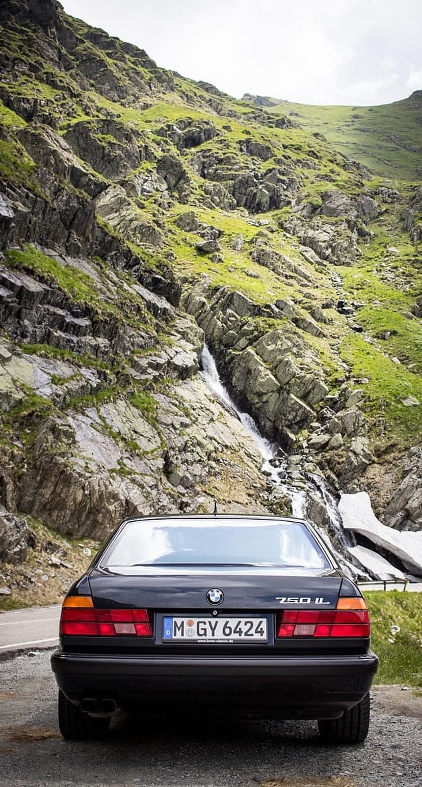 Black BMW 750iL luxury sedan parked on a winding asphalt road next to a tall cascading mountain waterfall.