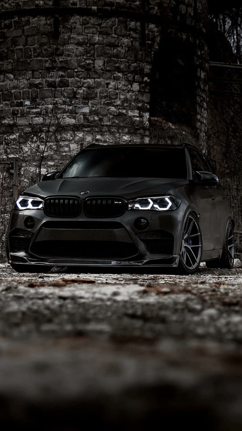 BMW X5 M SUV parked at night in front of a dark brick wall with illuminated LED headlights and nearby foliage.