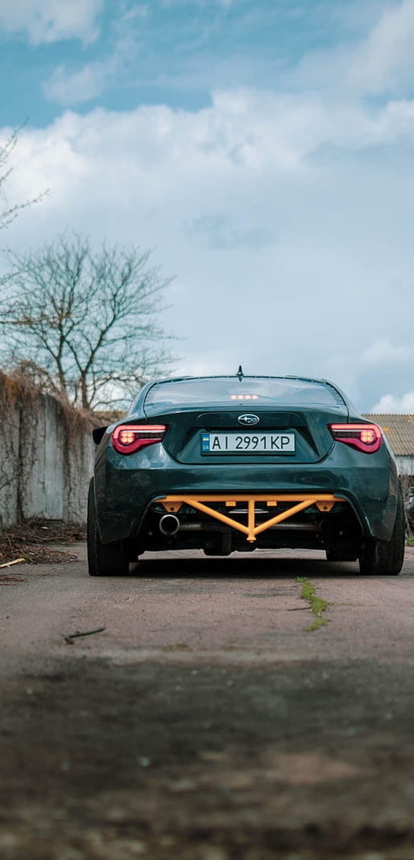 Subaru BRZ rear view showing red taillights and a yellow strut brace against an urban concrete wall background.
