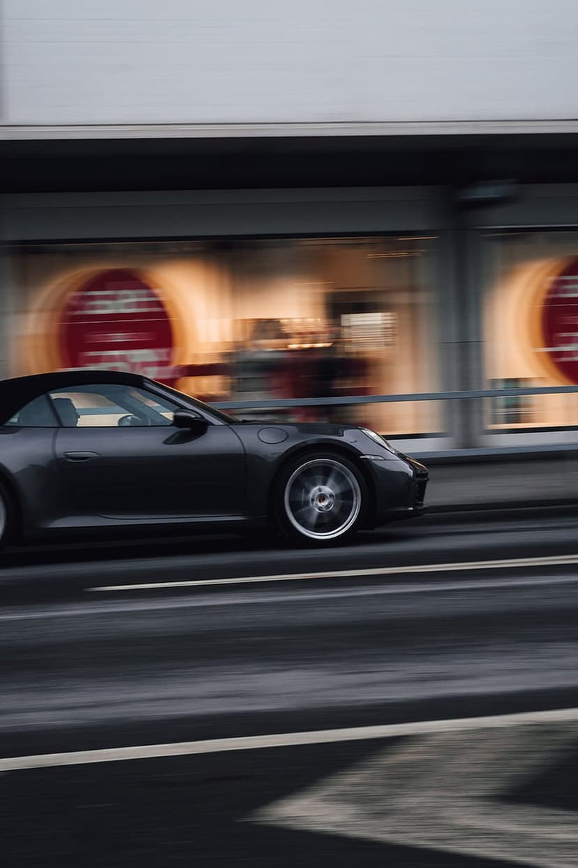 Dark Porsche convertible speeds past blurred red storefront signage on a wet asphalt road at night.