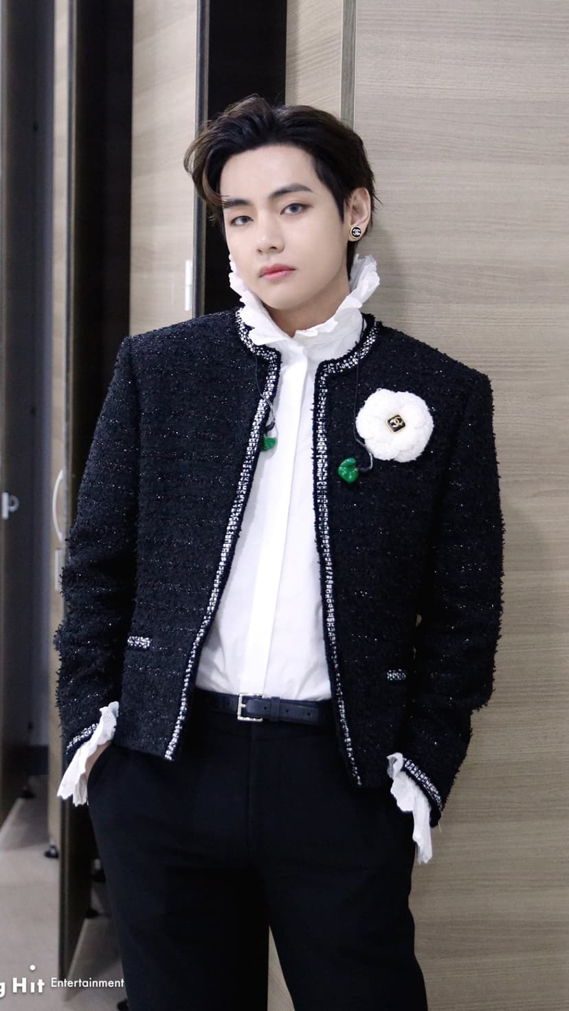 Kim Taehyung wearing a black tweed jacket with a white floral brooch and a ruffled white collar shirt.