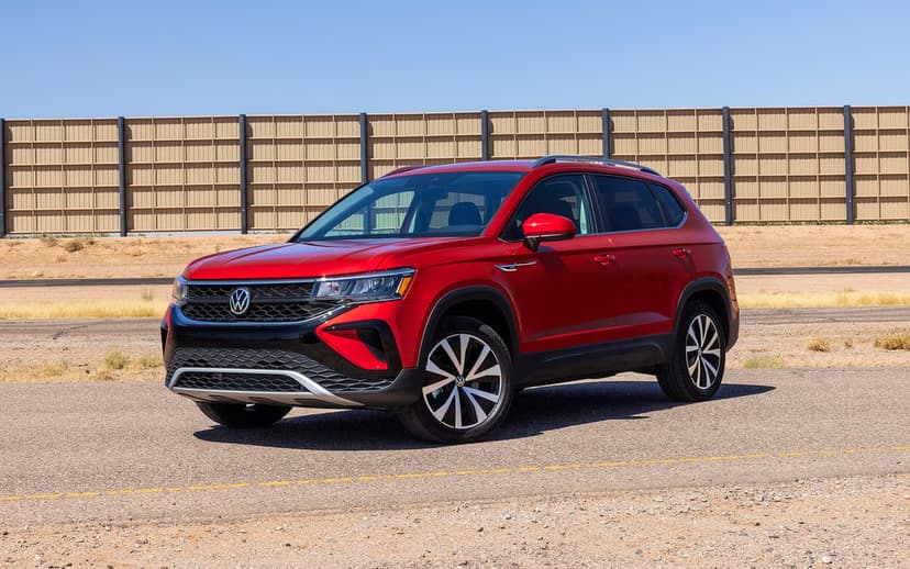 Red Volkswagen Taos SUV parked on an asphalt road with a tan industrial fence and clear blue sky background.