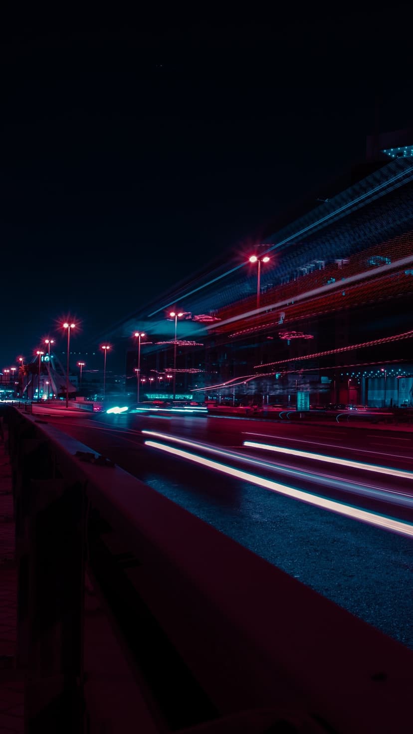 Red and white light trails curve along a dark highway under glowing blue and pink urban atmospheric lighting.