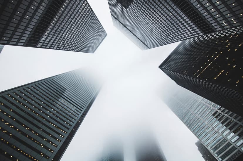 Dark modern skyscrapers stretch upward into a dense gray fog viewed from a dramatic low-angle perspective.