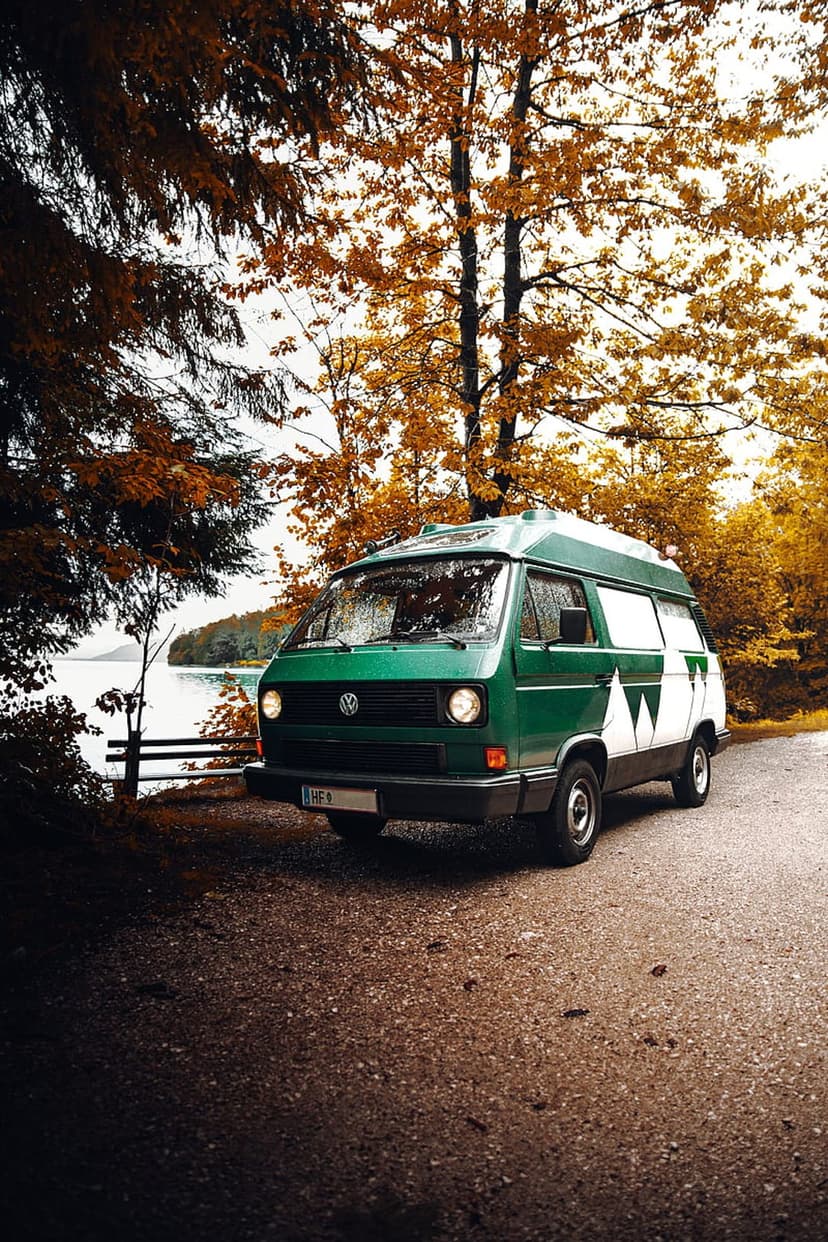 Green vintage camper van with white stripes parked on a gravel path next to a calm lake during peak autumn.
