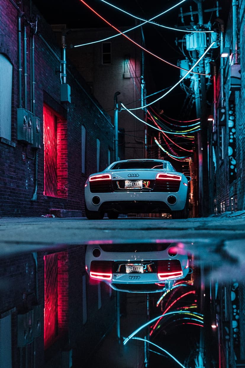 White Audi sports car in a dark alley reflecting in a wet puddle under glowing red and blue neon signage.