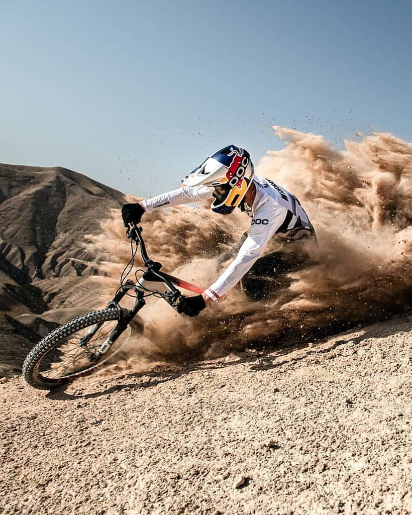 Mountain biker in full gear leans into a dusty turn on a steep desert slope under a bright blue summer sky.