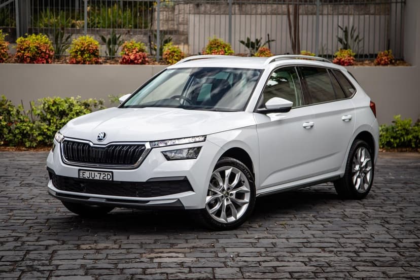 White Skoda Kamiq SUV parked on grey cobblestone pavement with a modern wall and green plants behind it.