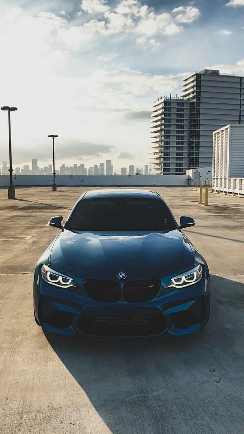 BMW M2 Coupe with glowing headlights parked in an urban lot during a dramatic sunset city skyline backdrop.