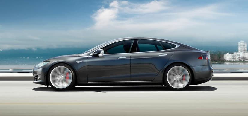 Charcoal grey Tesla Model S driving fast on a coastal highway with motion-blurred wheels and red brake calipers.