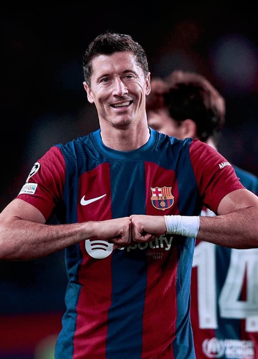 Robert Lewandowski smiles while performing a fist bump in a blue and red striped soccer jersey on a grassy field.