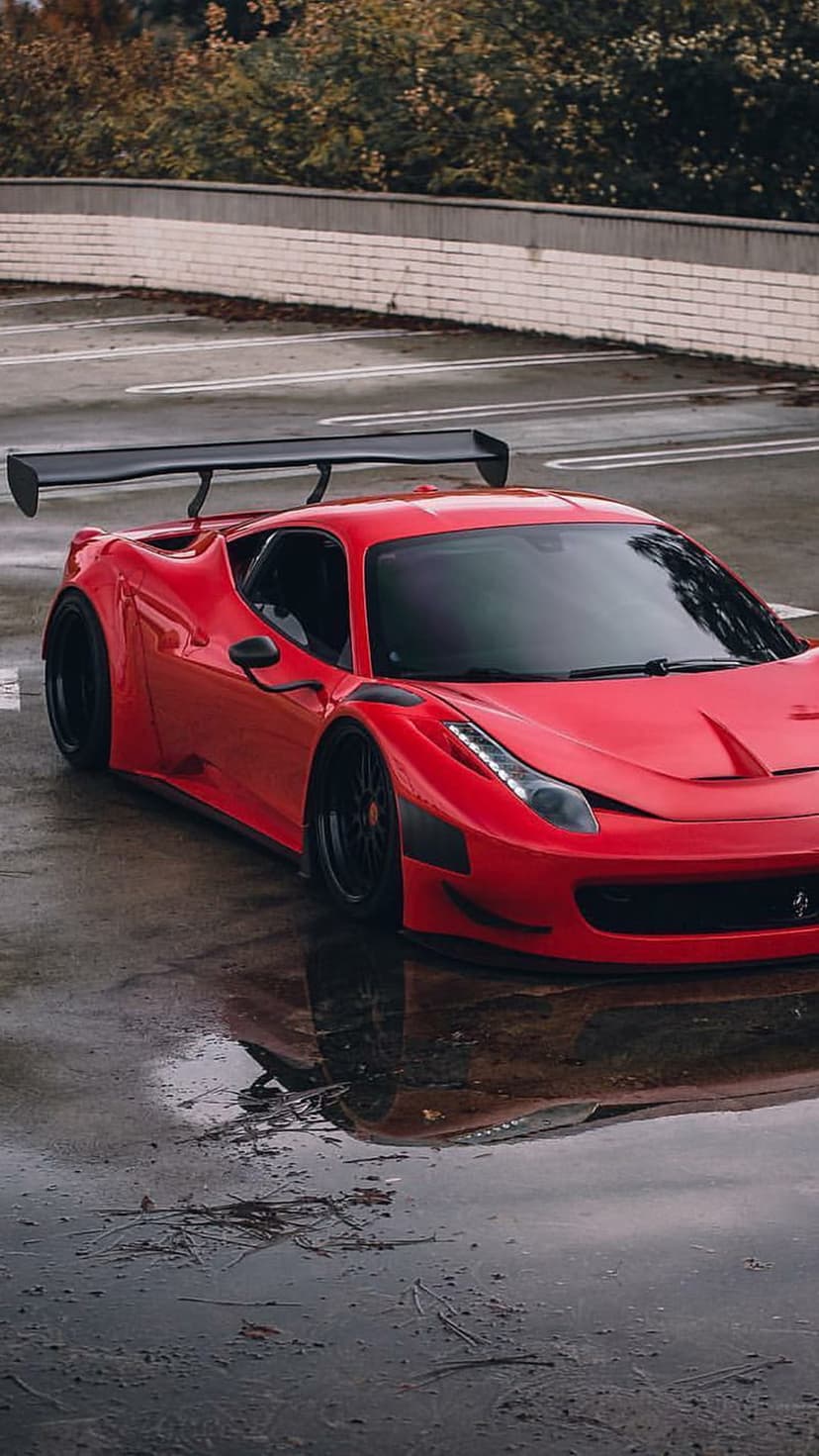 Red Ferrari 458 Italia with a large GT wing sits on wet pavement with reflections near a brick wall and trees.