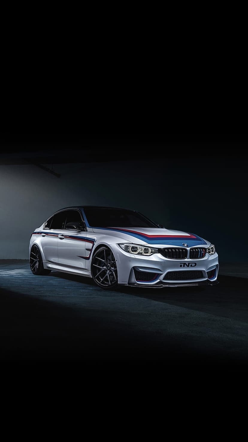 Silver BMW M3 sedan with M stripes and black rims parked in a dark studio with dramatic low-key lighting.