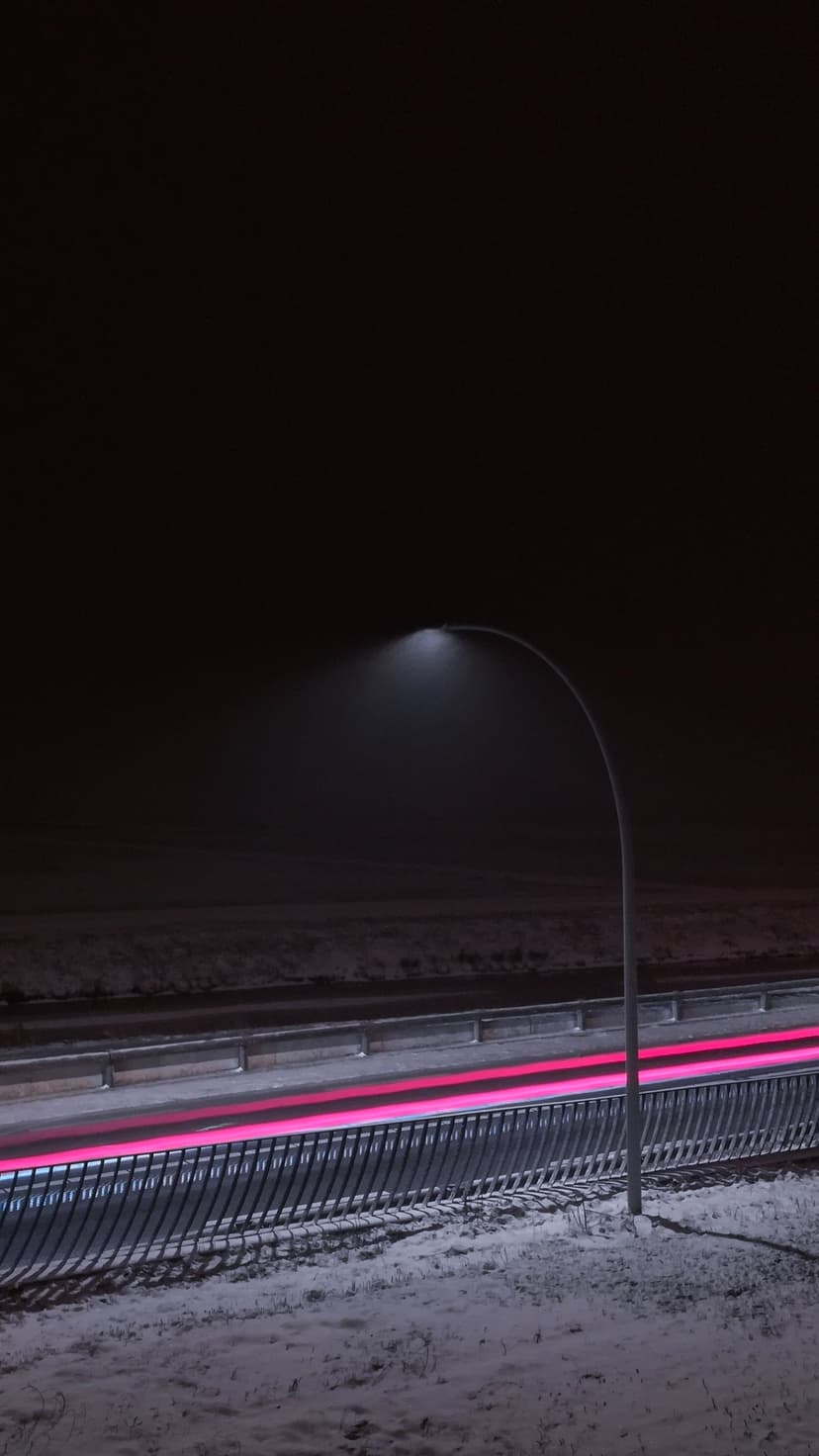 Red vehicle light trails curve along a snow-covered highway at night beneath a single glowing street lamp.