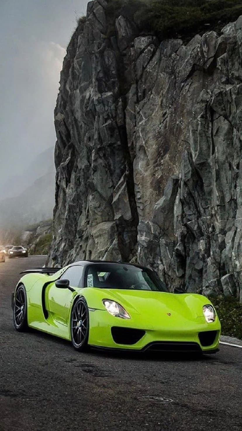 Green Porsche sports car driving on a winding mountain road near steep rocky cliffs under a foggy grey sky.