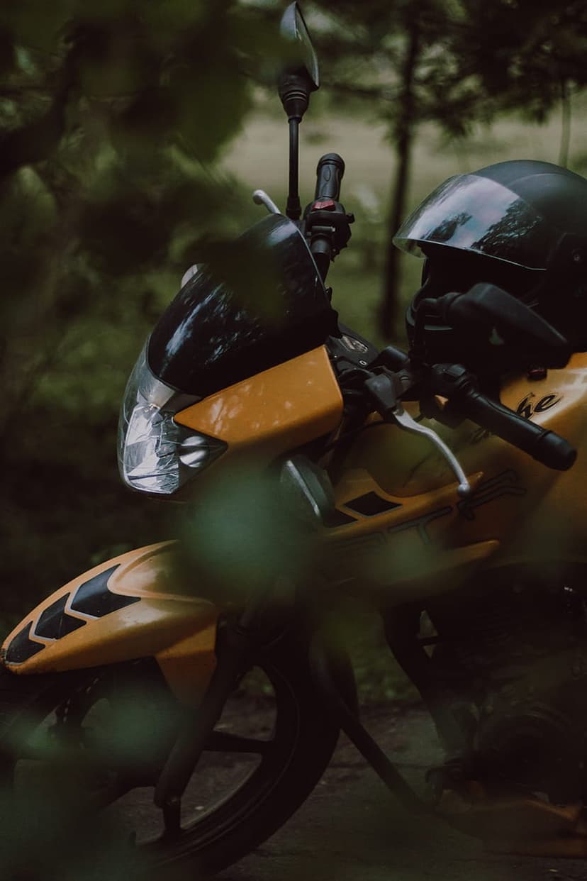 Yellow motorcycle with a black helmet parked on a dirt path in a dark, foggy forest with blurred green leaves.
